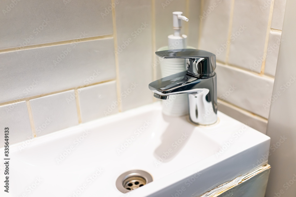 Modern bathroom stainless steel metal faucet in small tiny studio apartment sink closeup on tiles tiled wall of room and soap dispenser Modern bathroom stainless steel metal faucet in small tiny studio apartment sink closeup on tiles tiled wall of room and soap dispenser