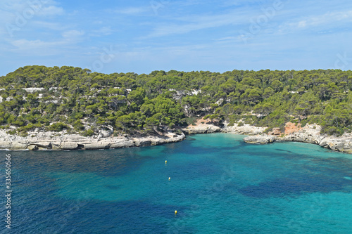 Fototapeta Naklejka Na Ścianę i Meble -  Tranquil summer landscape in Menorca, Spain. Turquoise sea, cliffs with pine trees, blue sky and light clouds.