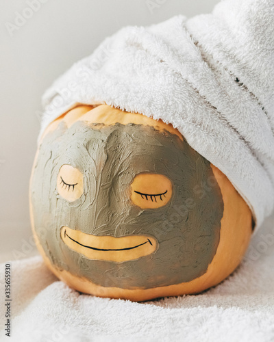 A cute smiling pumpkin with facial clay mask and white towel on light background. Spa, relax, health and beauty, face skin care concept. Resting