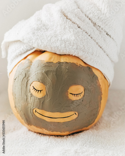 A cute smiling pumpkin with facial clay mask and white towel on light background. Spa, relax, health and beauty, skin care concept. Halloween