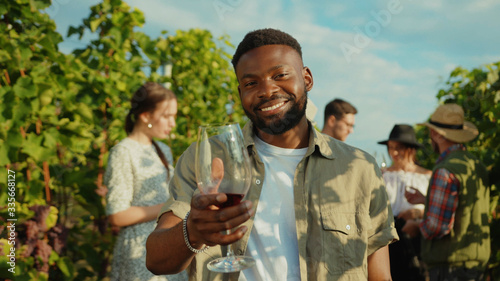 Smiling african young man holding wineglass toasting having a good time on wine tasting degustation holiday with friends. Vineyard.