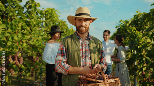 Portrait of proud caucasian farmer in strawhat smiling with joy holding large basket of grapes working during harvest in vineyard.