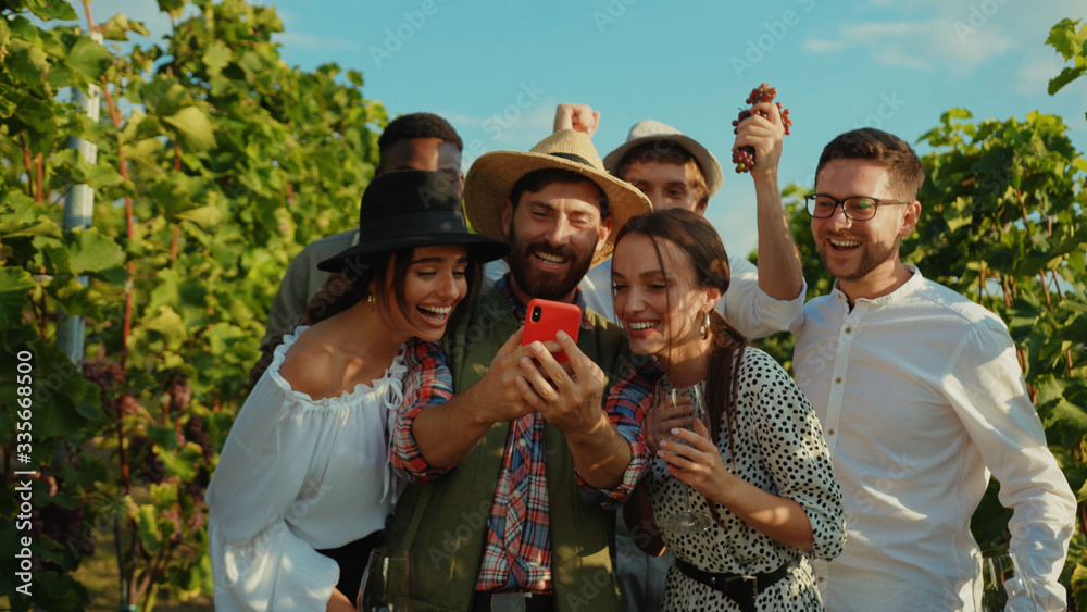 Group of attractive young friends laughing of fun content on smartphone hanging out staying at vineyard nature in summertime.