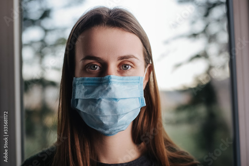 Close up portrait of young woman sitting on window sill at home  wearing virus mask  thinks about risk of epidemic disease  girl looks sadly at camera. Concept safety  coronavirus  virus protection