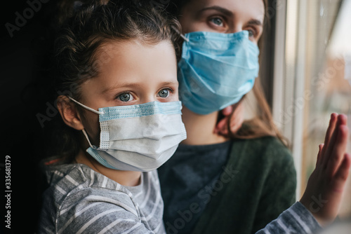 Unhappy young mother embracing upset little curly daughter with virus mask, sitting on windowsill at home, consoling sad preschool girl. Concept of coronavirus or COVID-19 pandemic disease symptoms