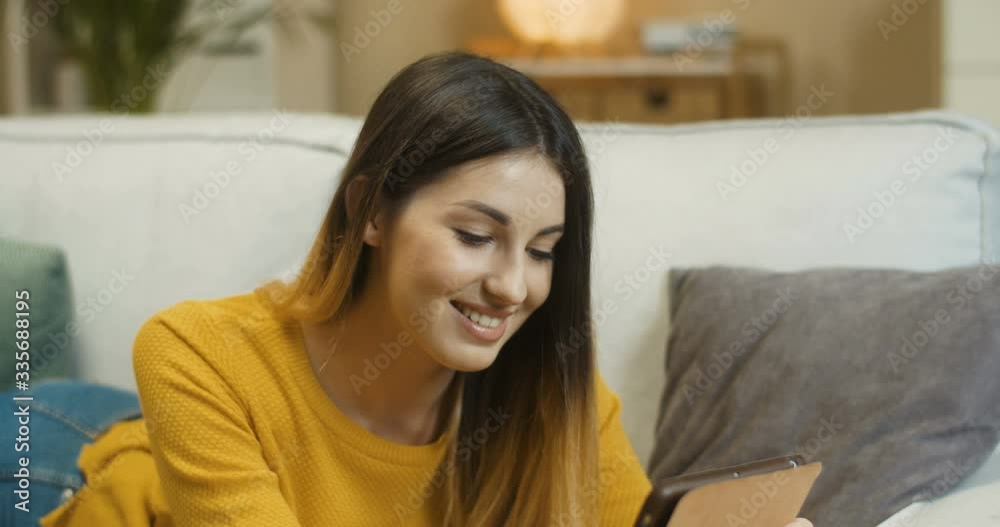 Close up portrait of happy smiling beautiful girl typing on tablet while lying on sofa in living room. Cheerful pretty Caucasian young woman texting on device at home in quarantine. Leisure concept Close up portrait of happy smiling beautiful girl typing on tablet while lying on sofa in living room. Cheerful pretty Caucasian young woman texting on device at home in quarantine. Leisure concept