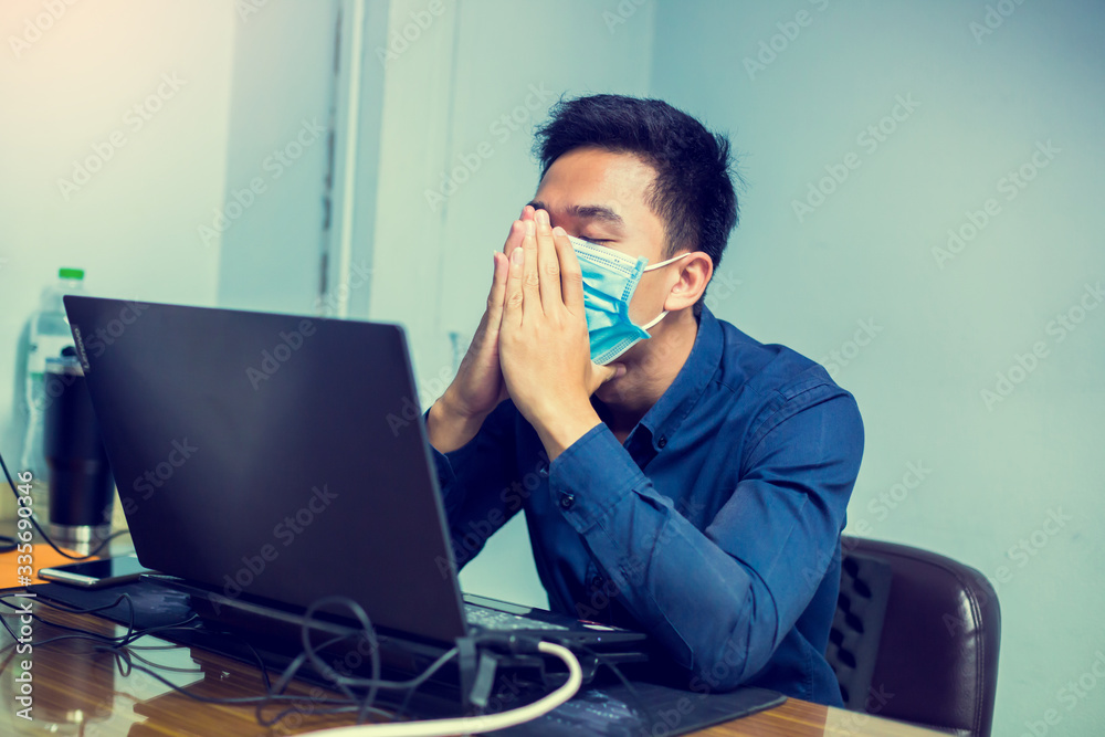 Businessman working with laptop. He wearing a mask and coughing in office. Asian male employee ill with Coronavirus Covid-19 infection. Concept of the spread of Coronavirus Covid-19 in office. Businessman working with laptop. He wearing a mask and coughing in office. Asian male employee ill with Coronavirus Covid-19 infection. Concept of the spread of Coronavirus Covid-19 in office.