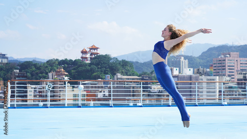 beautiful european girl dancer jumps high in the dance. curly sports model smiling on the background of the blue floor of a cruise liner and tropical views of Asian city and temple, mountains and sky
