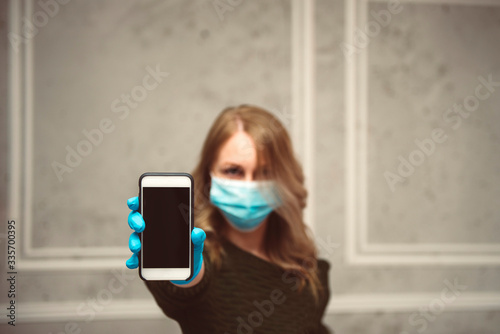 Girl in a medical mask and protective gloves holds a smartphone in her hand at home