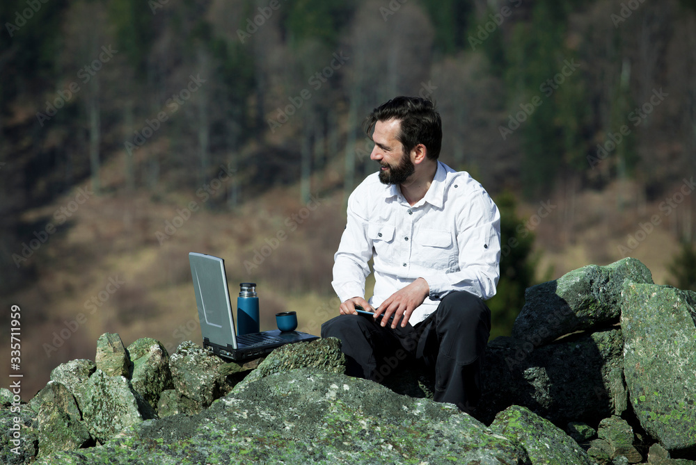 Active black bearded management worker business man in office shirt  working on laptop ant cell phone at mountain mossy rock at pine forest outback wasteland wild nature reach the top concept