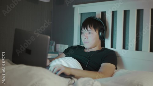 Young asian Man in headphones Using A Laptop Late At Night In Bed photo