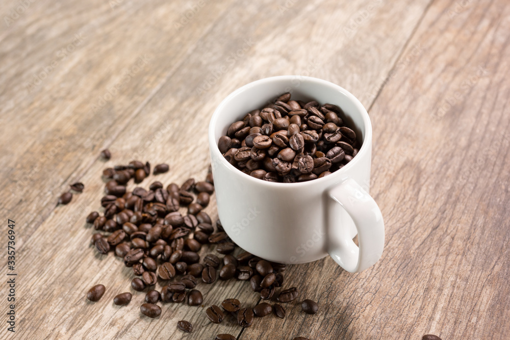 A view of a white mug filled with coffee beans, with a pile of coffee beans below in a mess on the table. A view of a white mug filled with coffee beans, with a pile of coffee beans below in a mess on the table.