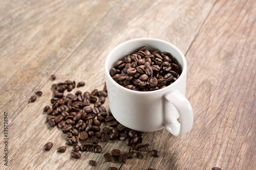 A view of a white mug filled with coffee beans, with a pile of coffee beans below in a mess on the table.