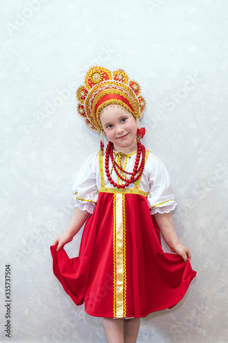 Child girl in Russian national costume sundress and kokoshnik. A child stands against a light wall. National traditions and culture