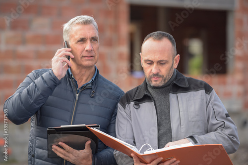 The architect telephones during the construction site  meeting while his contractor reads his folder