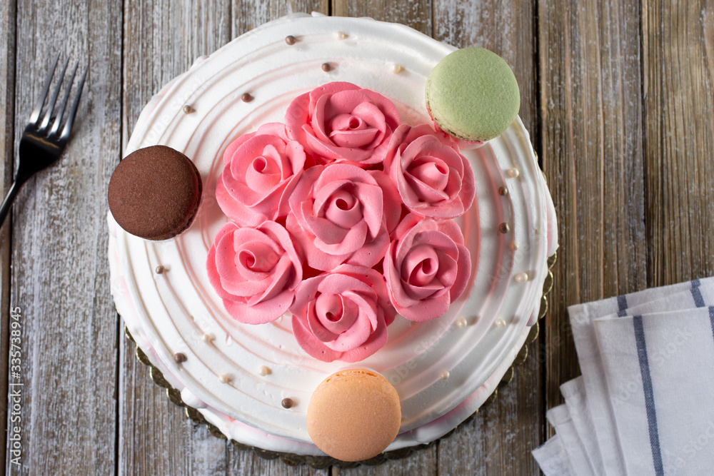 A top down view of a decorated cake with pink flower frosting designs and macaron cookies as toppings, in restaurant or kitchen setting. A top down view of a decorated cake with pink flower frosting designs and macaron cookies as toppings, in restaurant or kitchen setting.