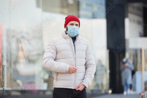A man with a beard wearing a medical face mask to avoid the spread coronavirus (COVID-19). A guy with a surgical mask on the face because of the pandemic zipping up a jacket in the center of the city. © Roman Tyukin