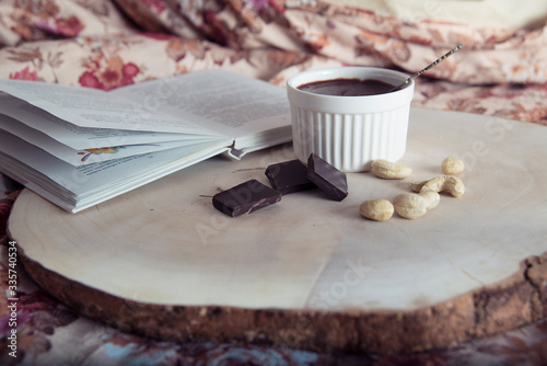 An open book, cashews, chocolate and pudding in a ceramic cup are on a wooden tray.
