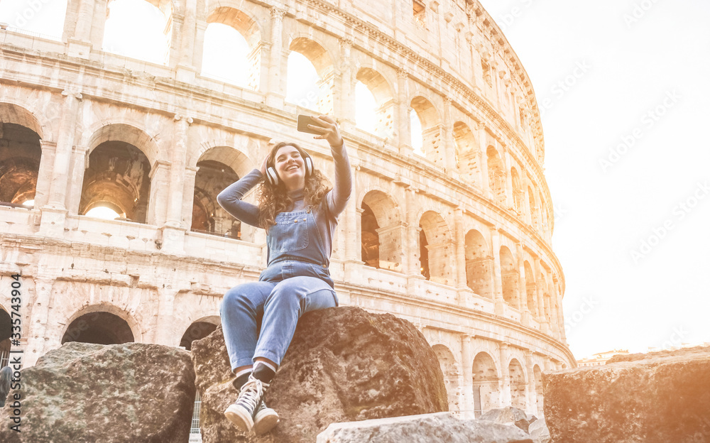 Young woman taking selfie in front of Coliseum in Rome while listening music with hedphones - Happy girl having fun with technology trends - Travel, real people and tech concept - Soft focus on face