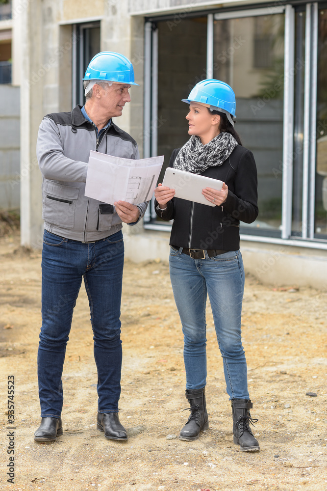 The worker reads the blue-print while the female architect is reading his notes on his tablet The worker reads the blue-print while the female architect is reading his notes on his tablet
