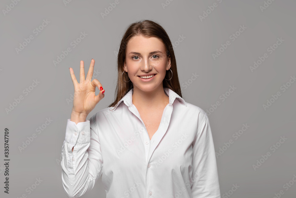 smiling young businesswoman in white shirt showing ok sign isolated on grey smiling young businesswoman in white shirt showing ok sign isolated on grey