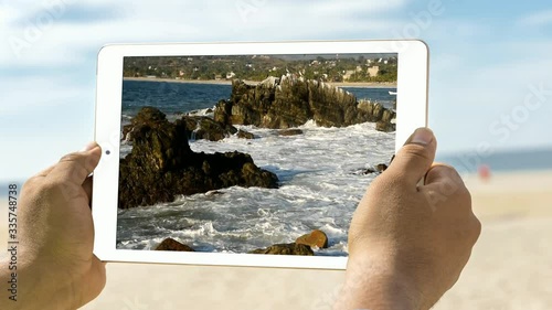 tablet held by two hands with video of rocks with pelicans on stormy sea, beach backgrounds, blue skies