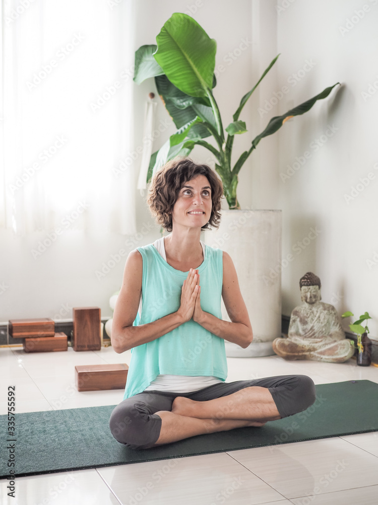 smiling woman with eyes open and hands in prayer position crossed legged half lotus on a yoga mat in a studio with cushions buddha and palm tree after meditation smiling woman with eyes open and hands in prayer position crossed legged half lotus on a yoga mat in a studio with cushions buddha and palm tree after meditation