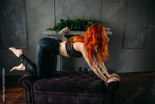 A sexy girl with long ginger hair in a black bra and leather pants is on all fours