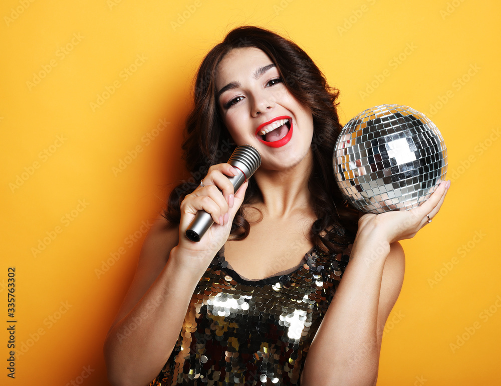 woman in evening dress holding microphone and disco ball over yellow background woman in evening dress holding microphone and disco ball over yellow background