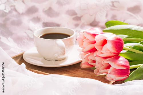 A cup of aromatic freshly ground coffee with a fresh bouquet of white-pink tulips on a wooden tray. Horizontal view