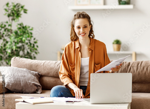 Fototapeta Happy dedicated woman examining documents and using laptop while working at home
