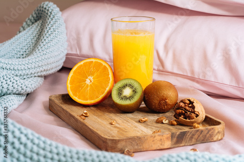 healthy and vegetarian breakfast in bed with freshly squeezed orange juice, half orange, sweet kiwi and walnut slices on a wooden tray against a backdrop of pink bedding, pillows and blue plaid