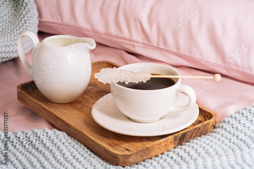 Breakfast in bed with freshly brewed and delicious coffee, a jug of cold milk and crystal sugar on a stick on a wooden tray against a backdrop of pink sheets, pillows and a blue plaid