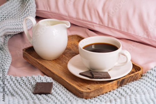 Breakfast in bed with freshly brewed and aromatic coffee  a jug of cold milk and three slices of real dark chocolate on a wooden tray against a backdrop of pink sheets  pillows and a blue plaid