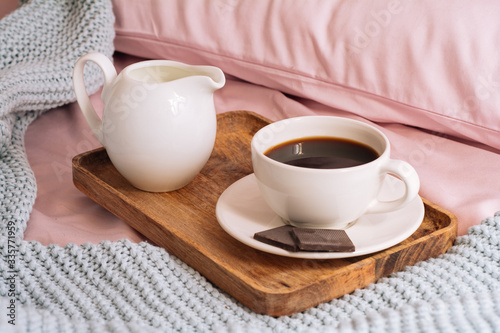 Breakfast in bed with freshly brewed and delicious coffee, a jug of cold milk and three slices of real dark chocolate on a wooden tray against a backdrop of pink sheets, pillows and a blue plaid
