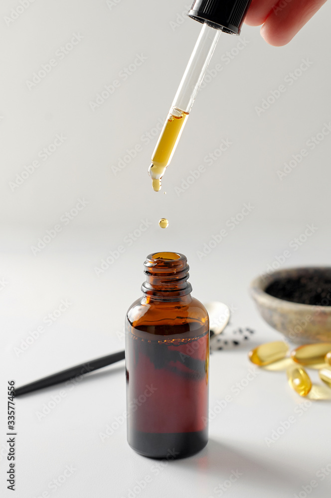 Pipette dropping essential black cumin seeds into a glass bottle. Nigella Sativa in spoon and bowl on white background