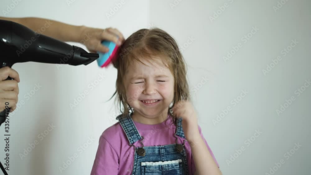 Closeup of mothers hands combing and blow-drying hair of little cute daughter. Little smiling girl closes eyes from hairdryer, child’s hair flying Closeup of mothers hands combing and blow-drying hair of little cute daughter. Little smiling girl closes eyes from hairdryer, child’s hair flying