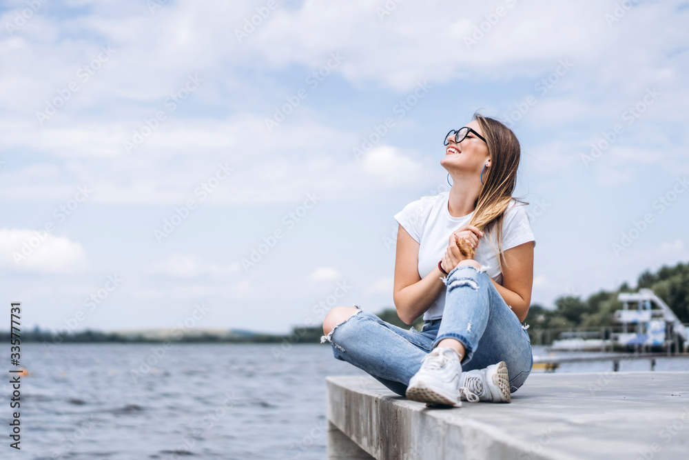 Young woman with long hair in stylish glasses posing on the concrete shore near the lake. Girl dressed in jeans and t-shirt smiling and looking away
