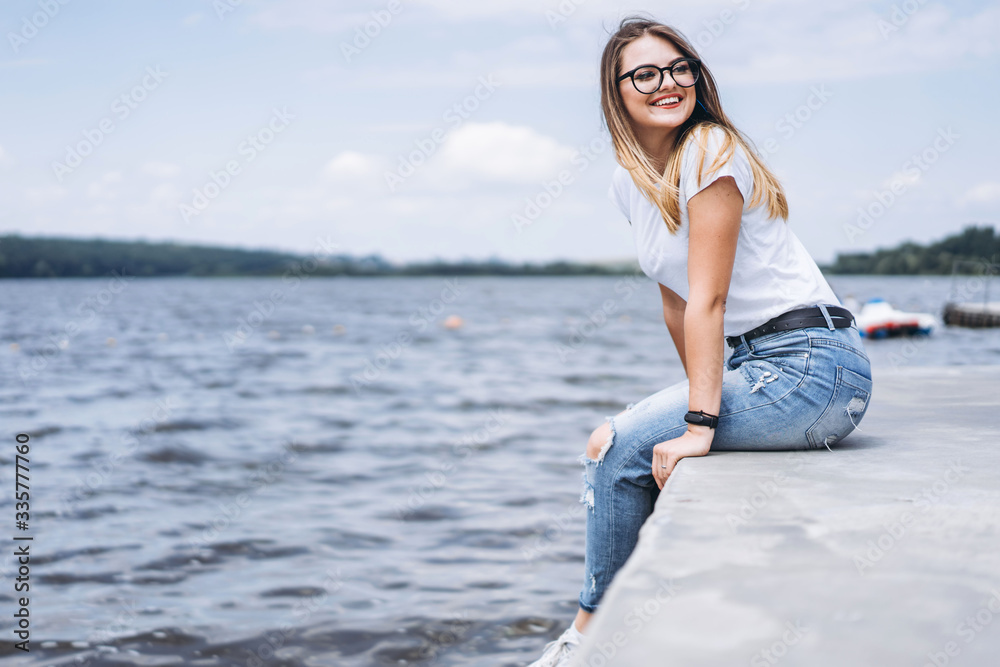 Young woman with long hair in stylish glasses posing on the concrete shore near the lake. Girl dressed in jeans and t-shirt smiling and looking away Young woman with long hair in stylish glasses posing on the concrete shore near the lake. Girl dressed in jeans and t-shirt smiling and looking away