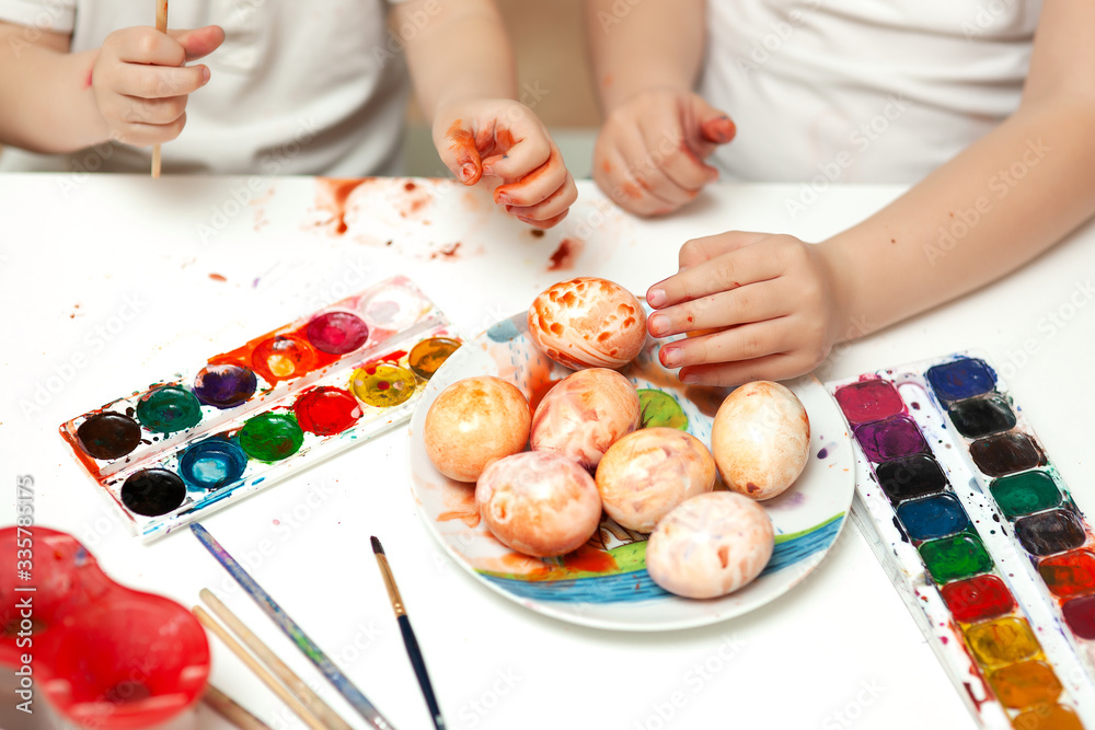 Bright watercolor paints lie on the table. Children's hands paint eggs. Easter
