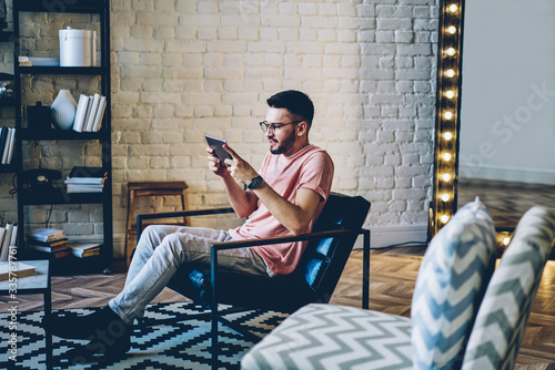 Young caucasian millennial man spending free time on online entertainment passing next level on portable pc at home interior, excited hipster guy playing games on digital tablet enjoying leisure photo