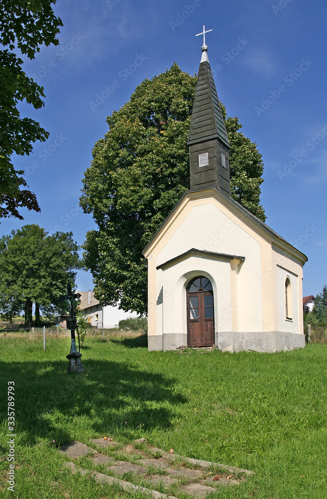 A small village chapel with a crucifix at the entrance against a background of green tree and blue sky.