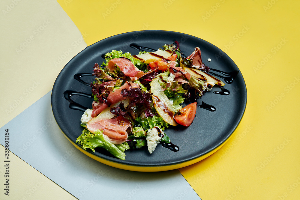 Tasty salad with lettuce, serrano jamon, pear, cherry tomatoes in black plate on colored background. Top view