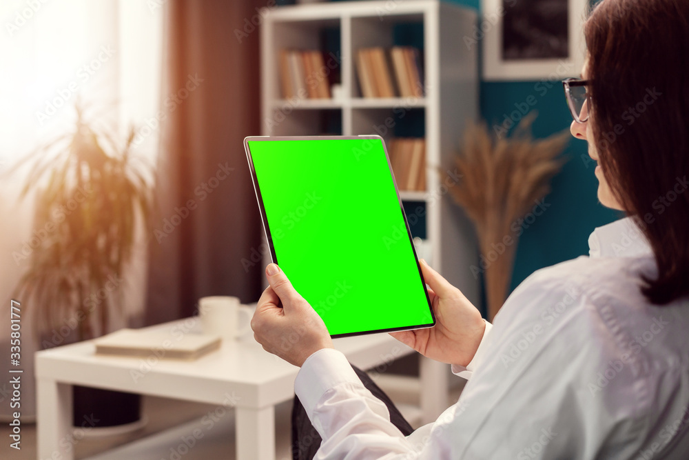 Over shoulder view of woman holding vertical tablet computer with green screen sitting in apartment - obrazy, fototapety, plakaty 