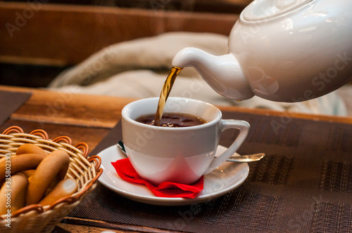Pouring fresh tea from porcelain tea kettle into white cup. Focus on tea stream, shallow depth of field.