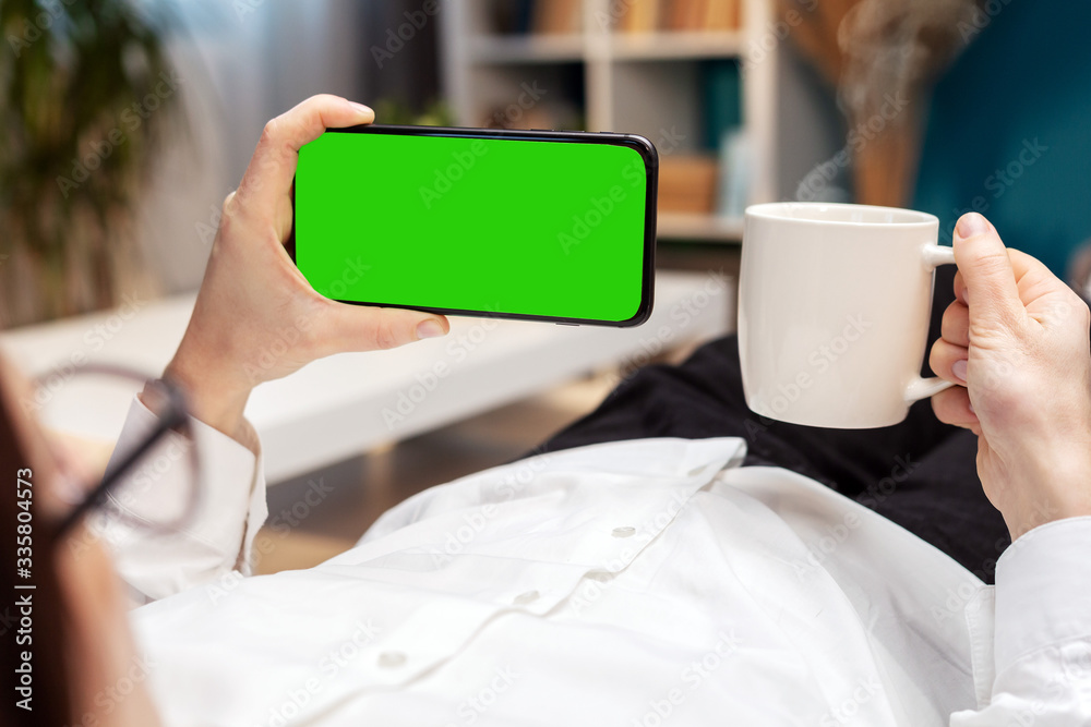 Back view of person holding smartphone in one hand and cup in the other lying on couch at home Back view of person holding smartphone in one hand and cup in the other lying on couch at home