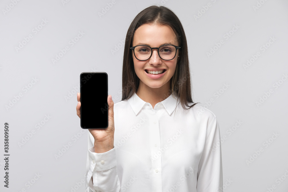 Young business woman wearing formal office shirt and big eyeglasses holding phone with blank screen and presenting it to viewer, copy space, isolated on gray background Young business woman wearing formal office shirt and big eyeglasses holding phone with blank screen and presenting it to viewer, copy space, isolated on gray background