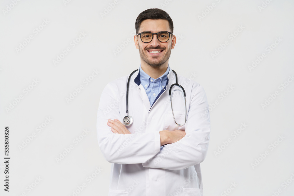 Portrait of smiling young male doctor with stethoscope around neck standing with crossed arms in white coat, isolated on gray background Portrait of smiling young male doctor with stethoscope around neck standing with crossed arms in white coat, isolated on gray background