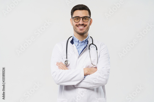 Portrait of smiling young male doctor with stethoscope around neck standing with crossed arms in white coat, isolated on gray background
