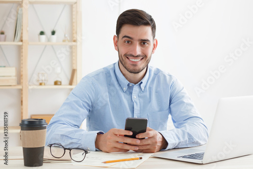 Young confident man spending lunch break in office, having rest from work while reading funny messages from friends and colleagues, smiling at camera